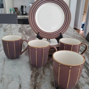 May include: Set of four purple mugs with gold accents and a white interior. The mugs are arranged on a table with a purple and white patterned plate in the background.