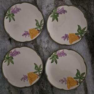 Puede incluir: Juego de cuatro platos de postre de cer&aacute;mica blanca con un dise&ntilde;o floral verde, amarillo y morado. Cada plato presenta una disposici&oacute;n diferente de las flores.