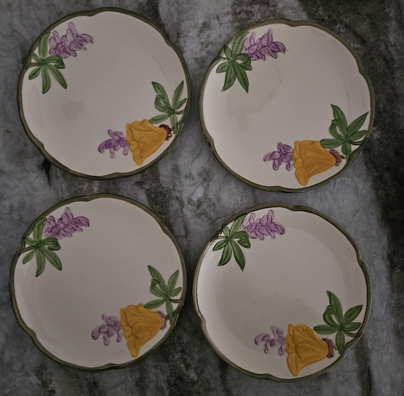 Puede incluir: Juego de cuatro platos de postre de cer&aacute;mica blanca con un dise&ntilde;o floral verde, amarillo y morado. Cada plato presenta una disposici&oacute;n diferente de las flores.