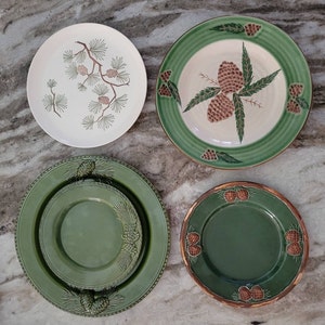Può includere: Quattro piatti decorativi con un design a pigna. Due piatti sono bianchi con pigne e foglie verdi. Due piatti sono verdi con pigne e foglie bianche.