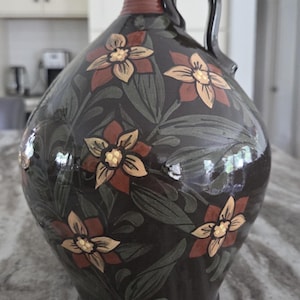May include: A dark brown ceramic jug with a glossy finish, featuring a hand-painted floral pattern in shades of muted green, rust orange, and cream. The vessel has a narrow neck, a single curved handle, and a rounded body with a wide base.