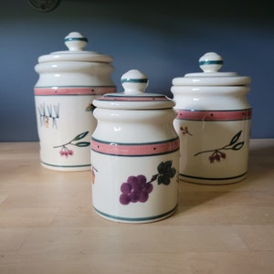 Puede incluir: Tres botes de cerámica con tapas, con diseños florales. Los botes son blancos con detalles en rosa y verde. El bote más grande tiene un diseño floral con una rama de agujas de pino. El bote del medio tiene un diseño floral con un racimo de uvas. El bote más pequeño tiene un diseño floral con una rama de bayas.
