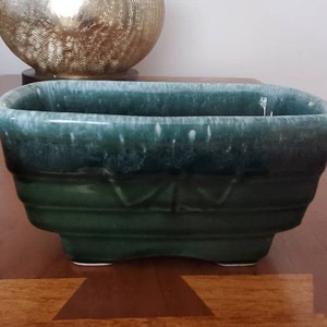 May include: A green glazed ceramic planter with a rectangular shape and a slightly rounded bottom. The planter has a white glaze on the top edge and a subtle pattern on the sides.