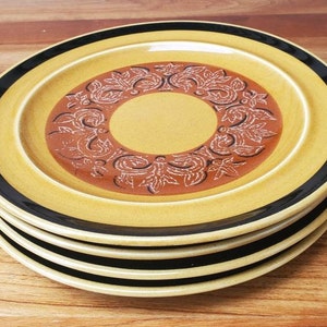 Pode incluir: Um conjunto de quatro pratos de jantar de cerâmica amarela com uma borda preta e um desenho floral castanho no centro.
