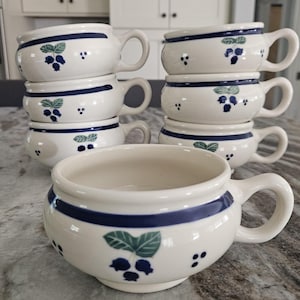 May include: A collection of white ceramic soup bowls with handles, adorned with a blue band and a blueberry and leaf motif. The bowls are stacked, displaying their design and form.