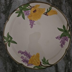 Puede incluir: Un plato de servir de cer&aacute;mica blanca con borde festoneado presenta un dise&ntilde;o floral de flores amarillas y flores de lupino p&uacute;rpura en un tallo verde.