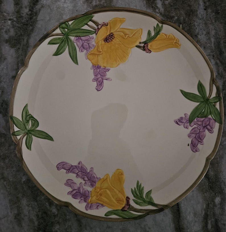 Puede incluir: Un plato de servir de cer&aacute;mica blanca con borde festoneado presenta un dise&ntilde;o floral de flores amarillas y flores de lupino p&uacute;rpura en un tallo verde.