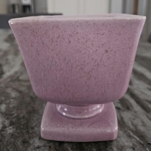 Könnte beinhalten: Ein lavendelfarbener Keramik-Pflanztopf mit gesprenkelter Textur. Der Pflanztopf hat einen quadratischen Sockel und eine ausgestellte, quadratisch geformte Oberseite. Der Sockel ist mit einem kreisförmigen Sockel mit der Oberseite verbunden.