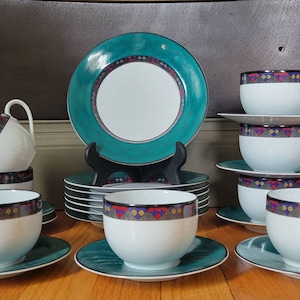 May include: A set of teal and white teacups and saucers with a geometric pattern. The cups and saucers are arranged in a circle on a wooden surface.
