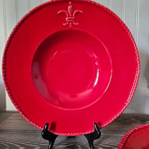 May include: A large, red ceramic soup bowl with a raised fleur-de-lis design. The bowl is on a black stand.