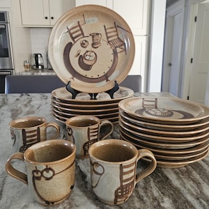 May include: A stoneware dinnerware set featuring plates and mugs with a hand-painted design of a table setting. The plates and mugs are in shades of tan and brown, with a rustic, artisanal aesthetic. The set includes multiple plates and mugs.