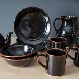 Könnte beinhalten: Ein Set aus braunen Tassen, Schalen und Krügen aus Keramik. Das Set beinhaltet eine Teekanne, einen großen Krug, einen kleineren Krug und sechs Tassen. Die Keramik ist komplett in einem dunklen Braunton glasiert mit einem leichten Glanz.