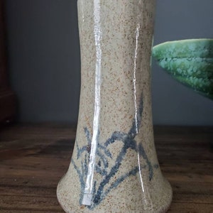 May include: A small, beige ceramic vase with a flared base and a blue floral design. The vase has a speckled glaze and a glossy finish.