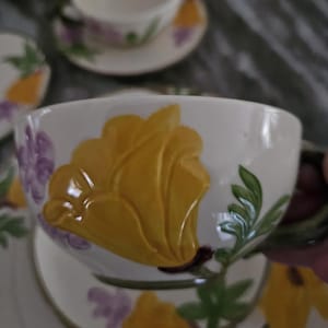 Puede incluir: Una taza de t&eacute; y un platillo blancos con un dise&ntilde;o floral. La taza presenta una gran flor amarilla con acentos morados. El platillo tiene un dise&ntilde;o floral similar.