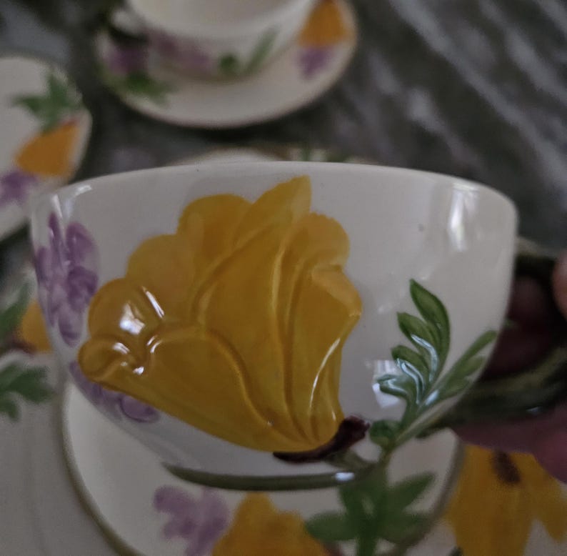 Puede incluir: Una taza de t&eacute; y un platillo blancos con un dise&ntilde;o floral. La taza presenta una gran flor amarilla con acentos morados. El platillo tiene un dise&ntilde;o floral similar.