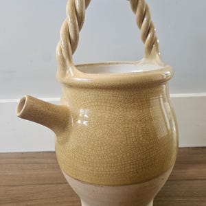 Puede incluir: Jarra de cerámica beige con asa trenzada y boquilla. La jarra tiene un esmalte craquelado y una base beige más clara. El asa es un diseño de cuerda trenzada. La jarra probablemente se utiliza para verter líquidos.