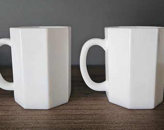 May include: Two white octagonal mugs with handles. The mugs are sitting on a wooden surface.
