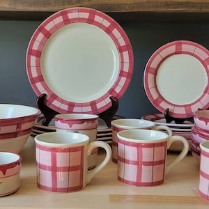 May include: A set of pink and white checkered ceramic dinnerware, including a large bowl, a smaller bowl, mugs, and plates. The set features a pink and white checkered pattern with a white background.