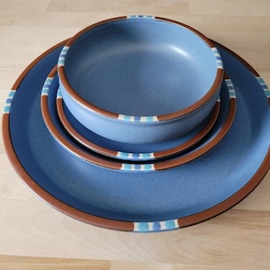 以下が含まれることがあります： 青いセラミック製の食器セット。大皿、小皿、ボウルが含まれています。縁は茶色で、青と白のストライプの装飾バンドが付いています。明るい木製の表面に配置されています。