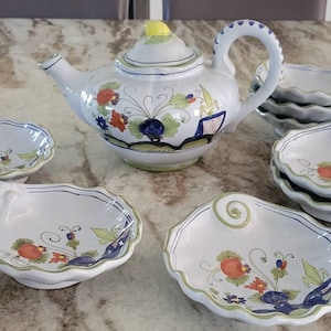 May include: A white teapot with floral designs in shades of blue, green, red, and yellow. The teapot is surrounded by six small white bowls with floral designs in similar colors. The bowls have a scalloped edge and a handle.