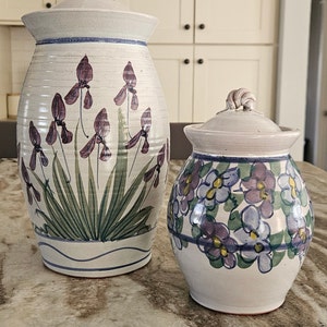 Può includere: Due vasi in ceramica con coperchi, entrambi decorati con motivi floreali dipinti a mano. Il vaso più grande presenta uno sfondo bianco con iris viola e foglie verdi. Il vaso più piccolo ha uno sfondo bianco con fiori viola e blu.