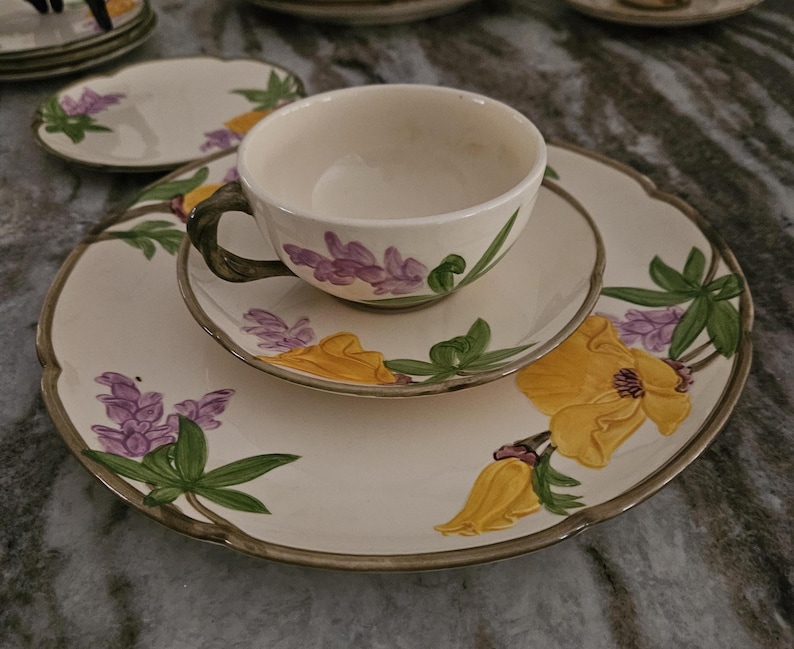 Puede incluir: Un juego de taza y platillo de t&eacute; blanco con un patr&oacute;n floral. El patr&oacute;n presenta flores amarillas, moradas y verdes sobre un fondo blanco.