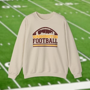May include: A cream-colored sweatshirt featuring a football-shaped design with a cityscape silhouette. The text "WASHINGTON FOOTBALL" is displayed in maroon and gold. The sweatshirt is set against a green football field background.