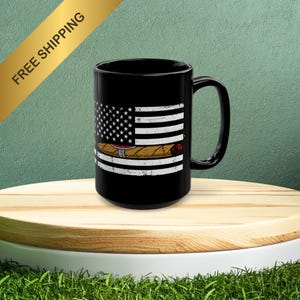 May include: Black ceramic mug with a white distressed American flag design and a cigar on top. The mug has a handle on the right side.