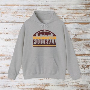 May include: A light grey hooded sweatshirt with a front pocket and a drawstring hood. The front displays a football design with the text "WASHINGTON FOOTBALL" in maroon and gold.
