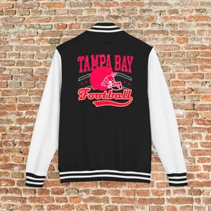 May include: Black and white varsity jacket with the text "TAMPA BAY Football" in pink and yellow. The design features a pink football helmet. The jacket has white sleeves and striped trim.