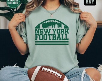 Könnte beinhalten: Hellgrünes T-Shirt mit dem grünen Schriftzug "NEW YORK FOOTBALL". Das Design zeigt eine Football-Form mit einer Skyline. Ein brauner und weißer Football liegt im Vordergrund.