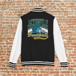May include: Black varsity jacket with white sleeves. The back features a teal football helmet graphic with the words "Jacksonville Football" in teal and gold. The jacket has a striped collar, cuffs, and hem.