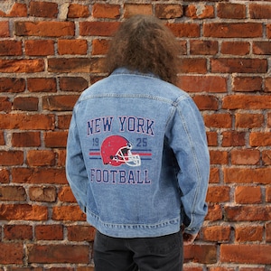 May include: A light blue denim jacket with a distressed look. The back of the jacket features a graphic with the words "NEW YORK FOOTBALL" and a red football helmet. The graphic includes the numbers "19" and "25".