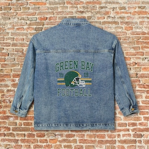May include: A light blue denim jacket with the words "GREEN BAY FOOTBALL" printed on the back. The design includes a green and yellow football helmet and the numbers "19" on either side of the helmet.