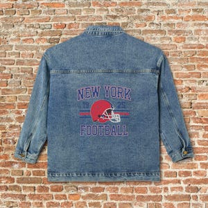 May include: A light blue denim jacket with a graphic on the back that says "NEW YORK FOOTBALL" with a red football helmet. The jacket has long sleeves, a button-up front, and two chest pockets. The jacket is made of a sturdy denim material.