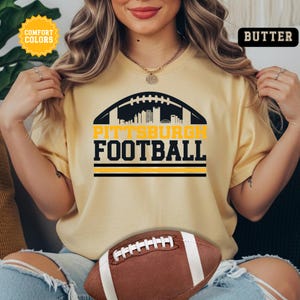 May include: A light yellow t-shirt with the words "PITTSBURGH FOOTBALL" in black and yellow, set against a football graphic. A brown football is in the foreground. The shirt has the words "COMFORT COLORS" and "BUTTER" on it.