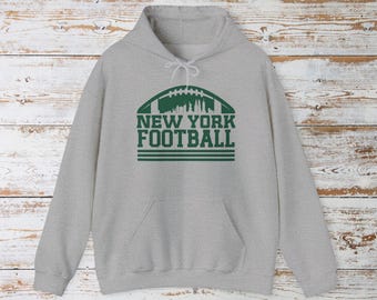 May include: A light grey hoodie with a front pocket and drawstring hood. The front features a green football graphic with the words "NEW YORK FOOTBALL" in a bold font. The hoodie is made of a soft, comfortable material.