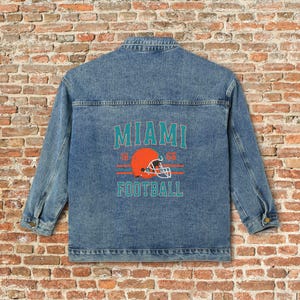 May include: Light blue denim jacket with a teal, orange, and white graphic on the back. The graphic reads "MIAMI FOOTBALL" with a football helmet design and the years "1966". The jacket has a classic button-up design.