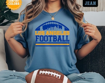 Könnte beinhalten: Blaues Jeans-T-Shirt mit den Worten "LOS ANGELES FOOTBALL" in Gelb und Blau. Das Design beinhaltet eine Footballform mit einer Skyline im Inneren. Ein brauner Football liegt im Vordergrund.
