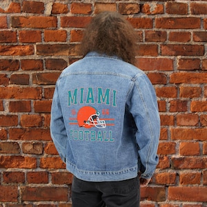 May include: Light blue denim jacket with the word "MIAMI" in teal block letters above an orange football helmet graphic. The word "FOOTBALL" is printed below the helmet in teal. The jacket has a classic button-up design.