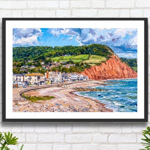 May include: A framed landscape painting depicting a coastal town nestled at the base of a large, reddish-brown cliff. The scene features a pebbled beach, blue ocean waves, and a hillside covered in green trees under a bright, cloudy blue sky.
