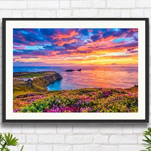May include: Framed landscape art print depicting a vibrant sunset over a coastal scene. The sky is filled with shades of blue, orange, and pink. The foreground features colorful wildflowers, with a cliffside and ocean in the background.