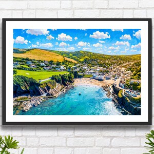 May include: A framed print depicting a coastal scene with a turquoise bay, sandy beach, and a town set amongst green hills. The sky is a vibrant blue with white clouds. The artwork is presented in a black frame.