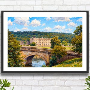 May include: A framed landscape painting depicts a stately building with a bridge over a river. The artwork features a stone bridge, lush green trees, and a blue sky with fluffy white clouds. The painting is in a black frame.