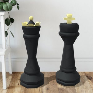 Puede incluir: Dos grandes piezas de ajedrez negras, un rey y una reina, se encuentran sobre un suelo de madera. El rey tiene una cruz dorada en la parte superior y la reina tiene una corona dorada.