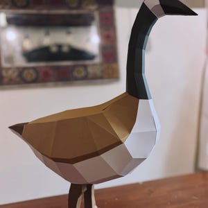 May include: A low-poly paper sculpture of a Canada goose featuring a geometric design. The goose has a black head and neck, a white throat patch, a brown body, and light brown legs. The structure is composed of flat, angular paper panels.