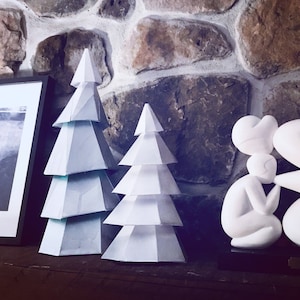 May include: Two white geometric Christmas trees with a layered design. The trees are displayed on a dark surface in front of a stone wall. A framed picture and a white sculpture are also visible.