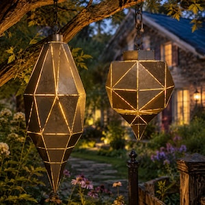 Op de afbeelding: Twee geometrische hanglantaarns gemaakt van verweerd grijs metaal met een gefacetteerd, meerzijdig ontwerp. Elke lantaarn heeft dunne, gloeiende lichtlijnen langs de naden en hangt aan donkere metalen kettingen aan een boomtak.