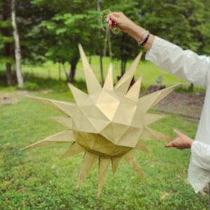 Może przedstawiać: Duża, złota papierowa latarnia w kształcie gwiazdy z geometrycznym wzorem. Latarnia jest zawieszona na sznurku i jest trzymana przez rękę.