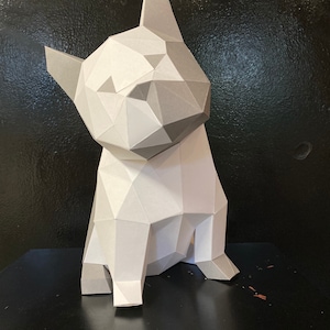 May include: A white and grey papercraft model of a French bulldog sitting on a black surface. The model is made of folded paper and has a geometric design.